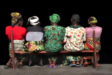Five African women sitting on a bench side-by-side.
