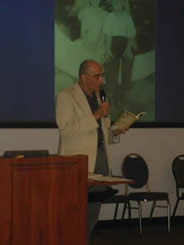 A man at a podium standing in front of a presentation, reading from his book.