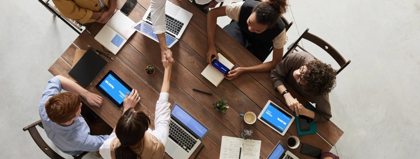 Overhead view of coworkers around a wooden table with laptops and tablets, two people shaking hands during a meeting.