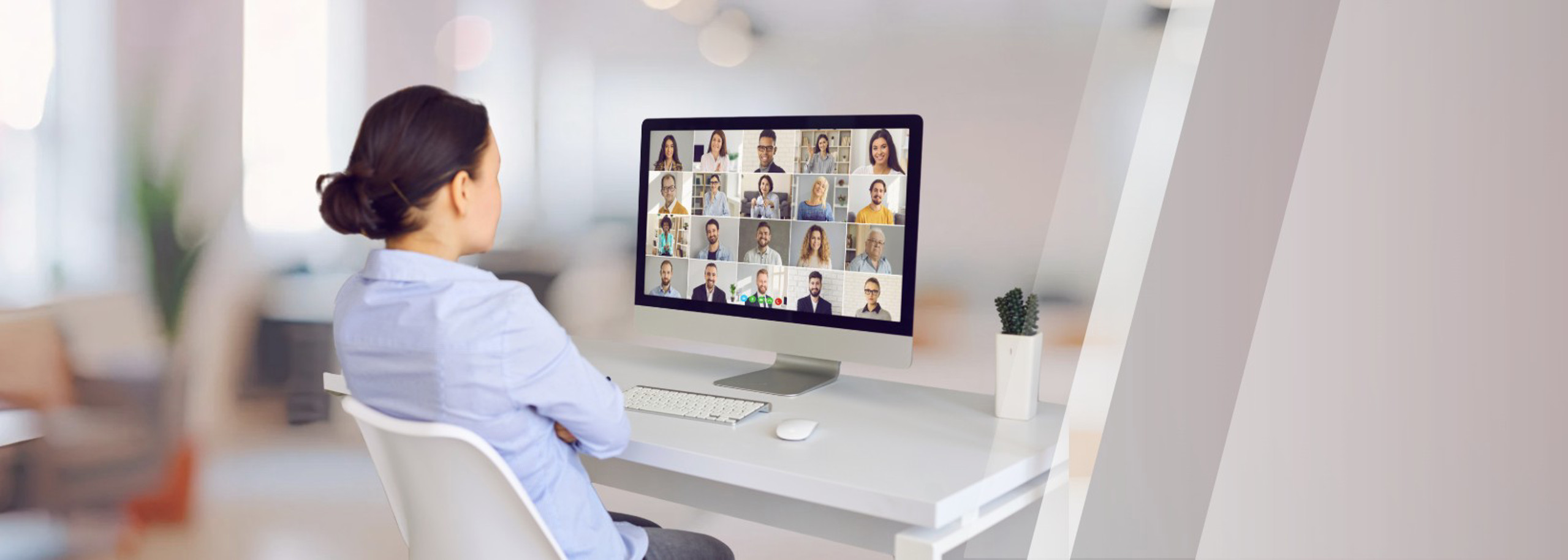 Person seated at a desk in a home office, viewing a video conference grid of multiple participants on a desktop computer monitor.