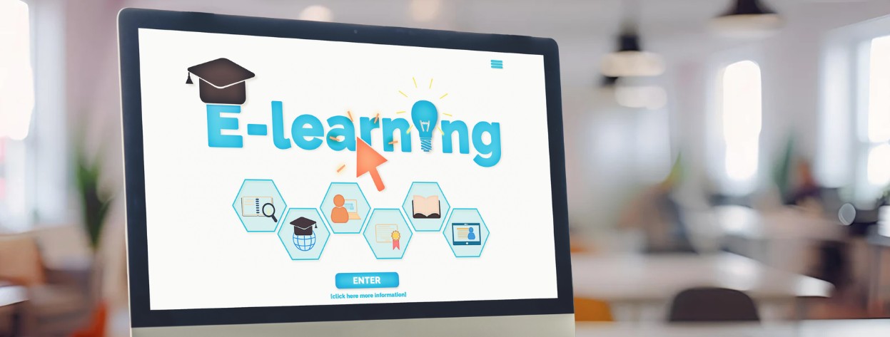 Computer monitor displaying an e-learning webpage with a graduation cap, lightbulb, and online education icons in a blurred office background.