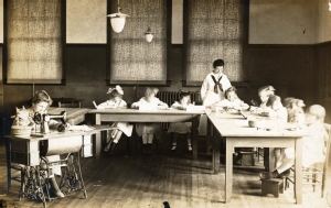 Photograph of old Training School classroom depicting a teacher showing young children to sew.