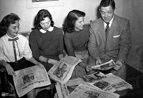 Three female student and a male faculty member look at newspapers.