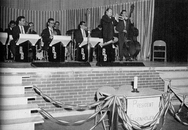 A band plays on stage at a VSC dance. An empty table with a sign for President Eisenhower in the foreground.