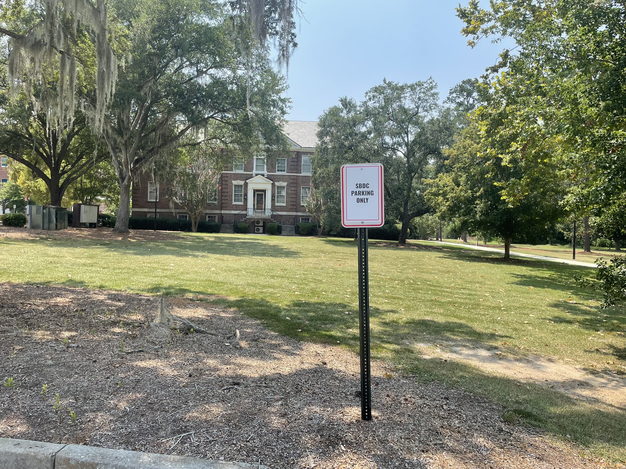 Sign reading “SBDC PARKING ONLY” in front of a brick building on a grassy, tree-lined campus.