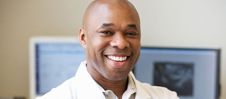 A smiling African American adult male wearing a white collared shirt is seen from the shoulders up. The background is blurred but two computer monitors are visible.