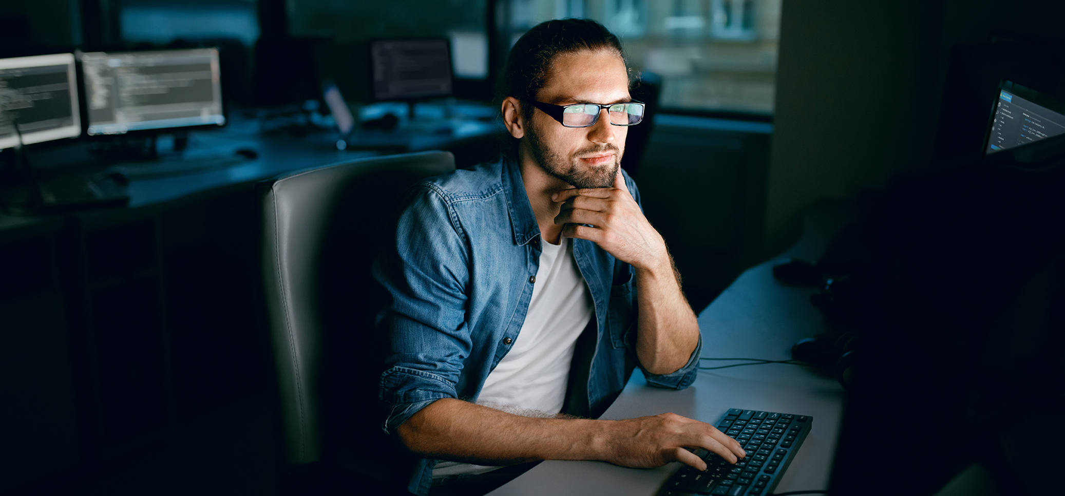 White male in his thirties with dark hair, beard, and glasses sits at a desk and stares thoughtfully at his computer monitor.  The background is blurred. Other computer monitors can be seen behind him on another desk area.
