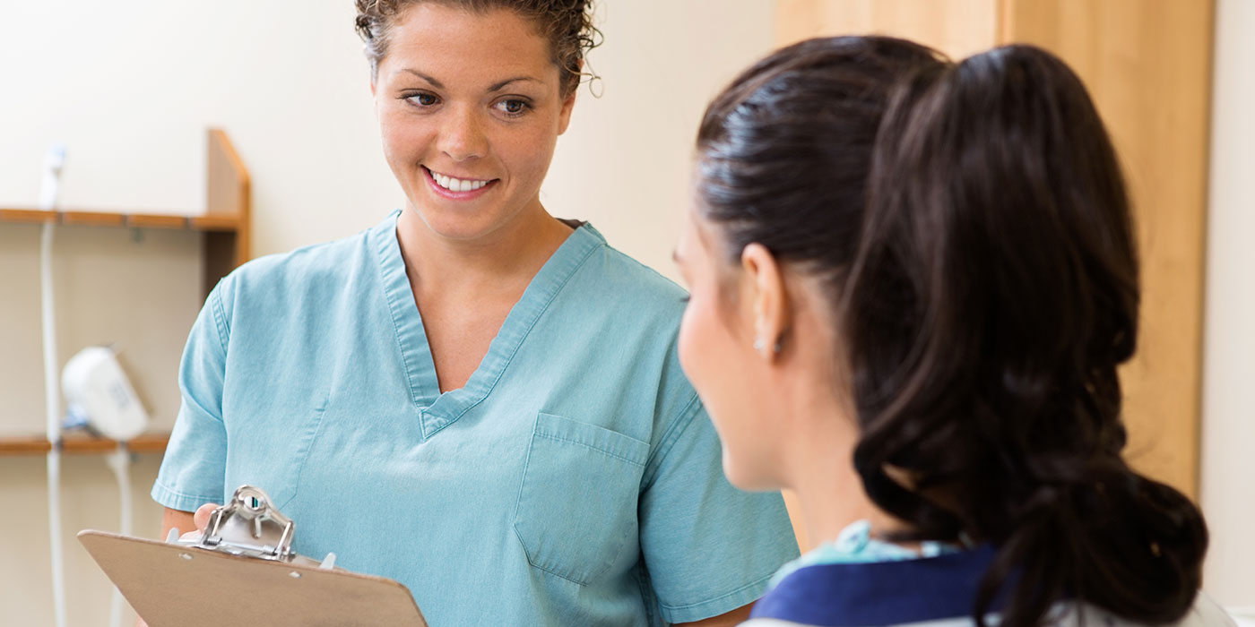 A young, smiling white woman with dark hair and wearing light blue scrubs is holding a clipboard. She is speaking to another young white woman with dark hair pulled up in a ponytail. The second woman's face is not visible because she is facing the first woman.