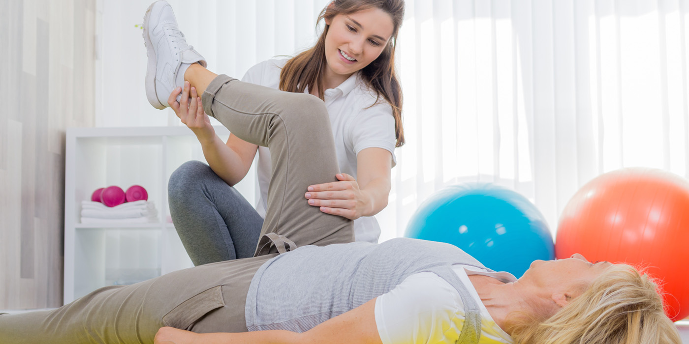 Physical therapy aide is exercising the leg of a female patient lying on a table.