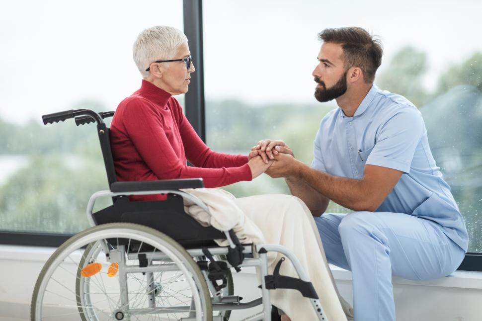 A white, older woman with white hair sits in a wheelchair. A white male healthcare worker is crouched in front of her, holding her hands in a supportive manner.