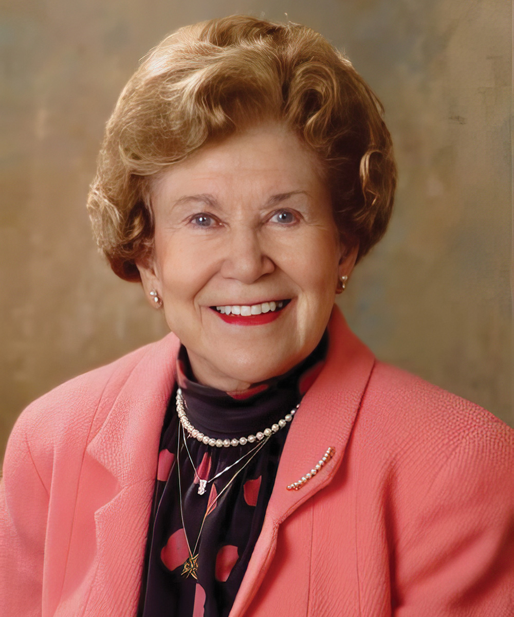 Smiling older woman in a pink blazer and patterned scarf, wearing pearl jewelry, posed against a neutral studio background.